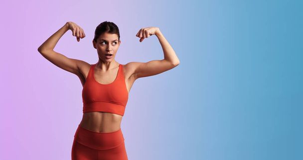 Young confident muscular female athlete with surprised face expression in tight orange activewear showing biceps while standing against gradient background - 写真・画像