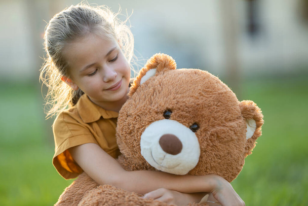 Pretty child girl tenderly embracing her teddy bear friend outdoors on green grass lawn. Concept of friendship values. - 写真・画像