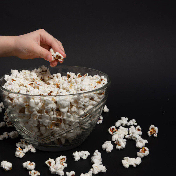 Un bambino prende popcorn da una ciotola, popcorn sparsi vicino a un piatto, sfondo nero e popcorn, spazio di copia e primo piano. - Foto, immagini