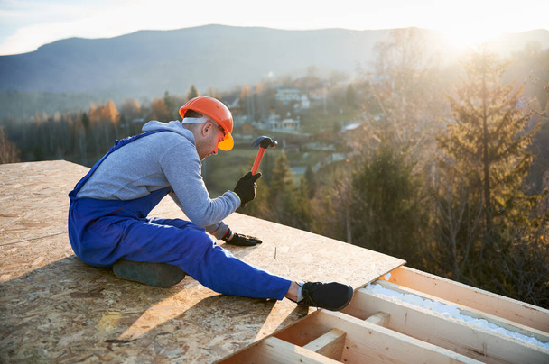Carpenter hammering nail into OSB panel on the roof top of future cottage in the evening. Man worker building wooden frame house. Carpentry and construction concept. - Фото, изображение
