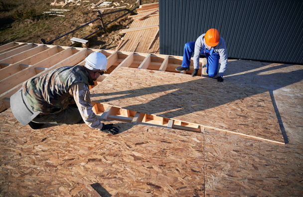 Carpenters mounting wooden OSB board on rooftop of future cottage. Men workers building wooden frame house. Carpentry and construction concept. - Photo, image