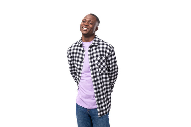 молодой красивый темнокожий мужчина в клетчатой рубашке и джинсах, улыбающийся в камеру на изолированном белом фоне с копировальным пространством. - Фото, изображение