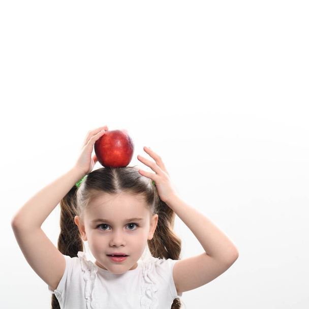 Egy vörös alma és egy kislány, egy fehér hátterű gyermek portréja, egy vörös alma a gyermek fején.. - Fotó, kép