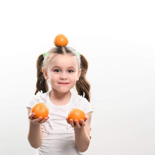 Portrait of a little beautiful girl on a white background with tangerines in her hands and one magdalene on her head, copy space and vitamin c. - Foto, immagini