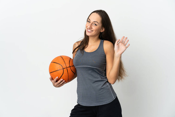 Mujer joven jugando baloncesto sobre fondo blanco aislado saludando con la mano con expresión feliz - Foto, Imagen