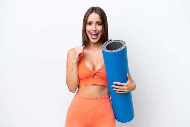 Young beautiful sport woman going to yoga classes while holding a mat isolated on white background celebrating a victory in winner position - Fotoğraf, Görsel