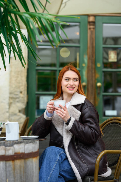 een roodharig meisje in een leren jasje. geweldige ochtend met koffie op het zomerterras in het café - Foto, afbeelding