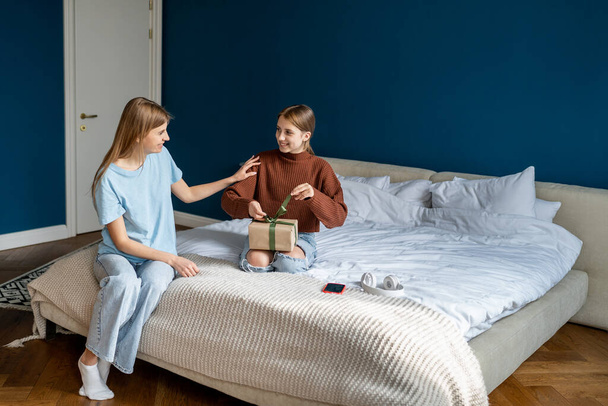 Mother congratulating teen girl daughter, giving wrapped gift box with ribbon. Caucasian family child and mom celebrate birthday at home together, adolescent kid sitting on bed opening present - Photo, Image