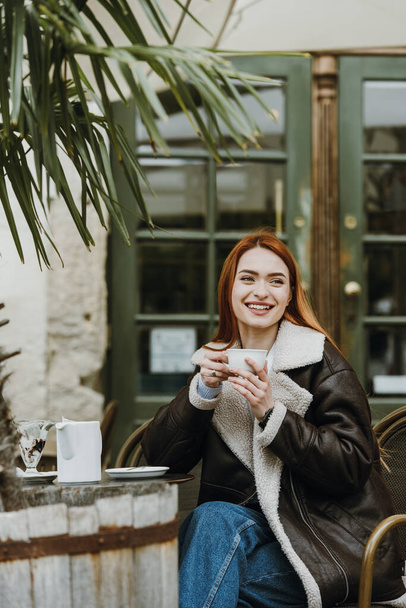 Rudowłosa dziewczyna w skórzanej kurtce. wielki poranek z kawą na letnim tarasie w kawiarni - Zdjęcie, obraz