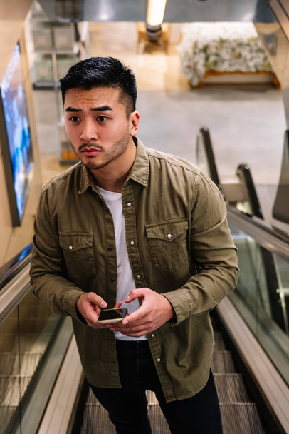 Ângulo alto do homem barbudo étnico adulto que navega o celular nas escadas móveis no centro comercial moderno durante o fim de semana - Foto, Imagem