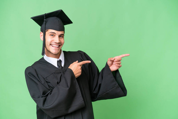 Giovane laureato uomo caucasico su sfondo isolato puntando il dito verso il lato e presentando un prodotto - Foto, immagini