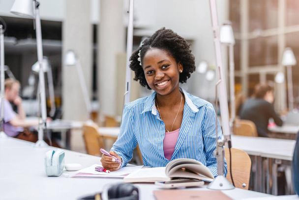 Egy fiatal fekete nő jegyzetfüzetbe ír egy könyvtárban. - Fotó, kép