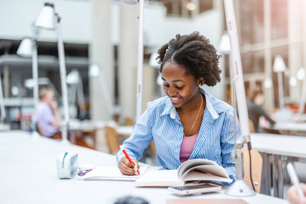 Jonge zwarte vrouw schrijven in notebook in een bibliotheek - Foto, afbeelding