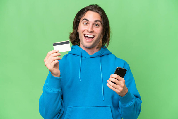 Fiatal jóképű férfi elszigetelt zöld króm háttér vásárol a mobil és a kezében egy hitelkártya meglepett kifejezés - Fotó, kép