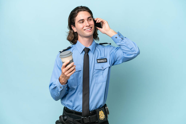 młody policjant biały człowiek odizolowany na niebieskim tle trzymając kawę do zabrania i telefon - Zdjęcie, obraz