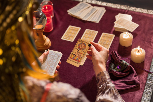 Korkea kulma sadon nimetön nainen ennustaja poiminta aurinko tarot kortti istuessaan pöydässä polttava kynttilöitä ja kristalli - Valokuva, kuva