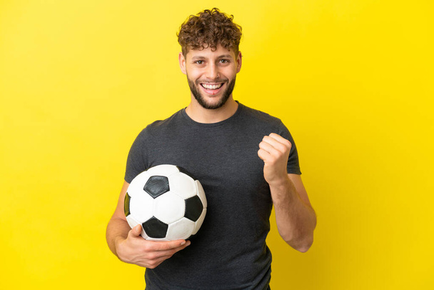 Όμορφος νεαρός ποδοσφαιριστής απομονωμένος σε κίτρινο φόντο γιορτάζει μια νίκη στη θέση του νικητή - Φωτογραφία, εικόνα