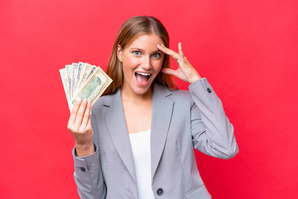 Junge kaukasische Geschäftsfrau hält Geld isoliert auf rotem Hintergrund mit überraschendem Ausdruck - Foto, Bild