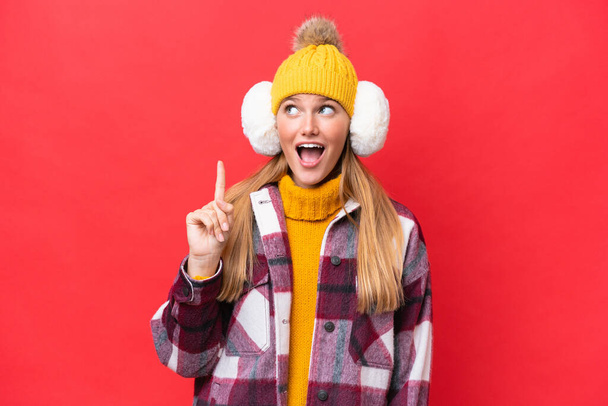 Young beautiful woman wearing winter muffs isolated on red background thinking an idea pointing the finger up - Photo, Image