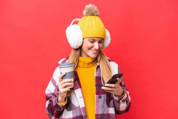 Jovem mulher bonita vestindo regalos de inverno isolado no fundo vermelho segurando café para levar e um celular - Foto, Imagem