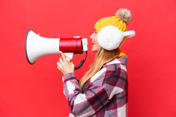 Young beautiful woman wearing winter muffs isolated on red background shouting through a megaphone - Foto, Imagen