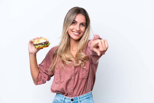 Młoda ładna urugwajska kobieta trzyma hamburgera odizolowanego na białym tle wskazuje palcem na ciebie z pewnym wyrażeniem - Zdjęcie, obraz