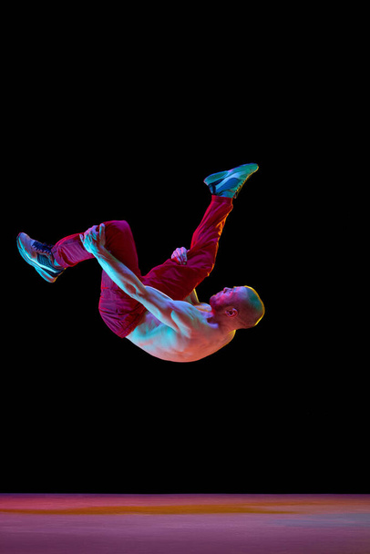 Somersault pose. Artistic young guy with strong muscular body dancing breakdance against black studio background in neon light. Concept of art, street style dance, fashion, youth, hobby, dynamics, ad - Foto, imagen