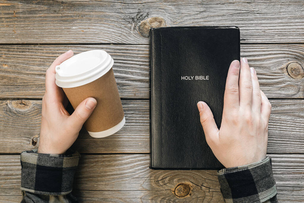 Bible and a paper cup of coffee in male hands on a wooden background, top view, concept of christianity and religion. - Zdjęcie, obraz