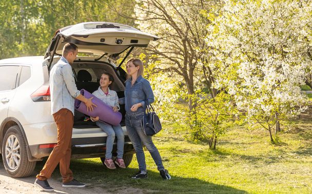Happy family standing together near a car with open trunk enjoying view of rural landscape nature. Parents and their kid leaning on vehicle luggage compartment. Weekend travel and holidays concept - 写真・画像
