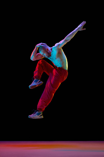 Young muscular man with relief shirtless body dancing breakdance against black studio background in neon light. Concept of art, street style dance, fashion, youth, hobby, dynamics, ad - Фото, изображение