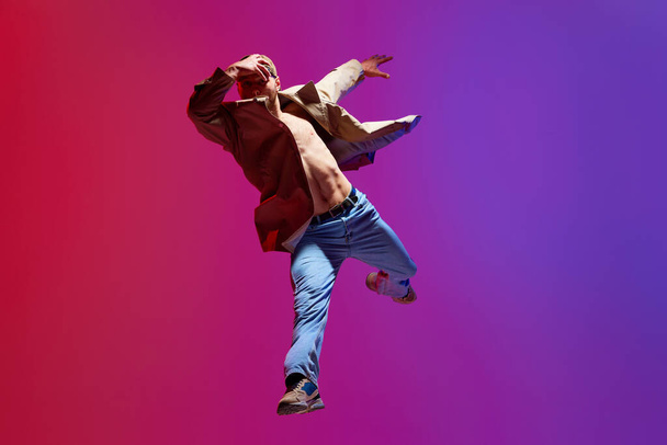 Active, artistic young man in casual clothes dancing breakdance, contempt against pink purple studio background. Concept of art, street style dance, fashion, youth, hobby, dynamics, ad - Photo, Image