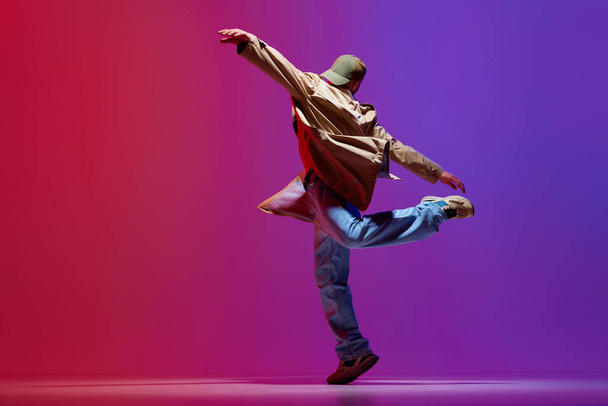 Dynamic image of young talented guy in stylish clothes dancing contemp, hip-hop against pink purple studio background. Concept of art, street style dance, fashion, youth, hobby, dynamics, ad - Fotó, kép