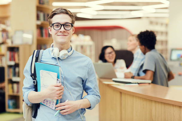 Portrait of smiling handsome young student in spectacles standing in public library and holding workbooks - Foto, afbeelding