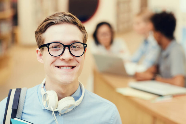 Portrait of smiling smart student boy in eyeglasses wearing headphones on neck spending time in library - Фото, изображение