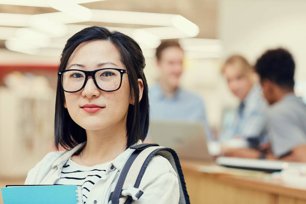 Portrait of confident intelligent Asian student girl in glasses wearing satchel standing in university hall, her groupmates chatting in background - Φωτογραφία, εικόνα