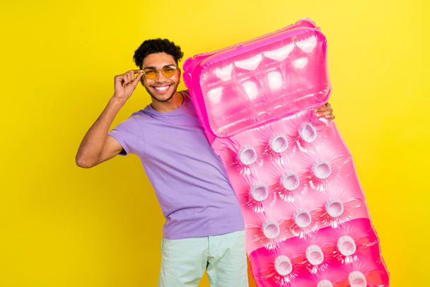 Portrait of carefree positive handsome man arm touch sunglass hold inflatable mattress isolated on yellow color background. - Fotó, kép