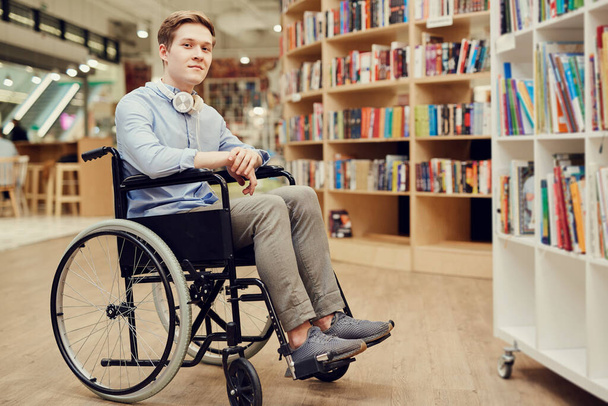 Portrait of content student boy with paralyzed legs sitting in wheelchair and choosing books in library - 写真・画像