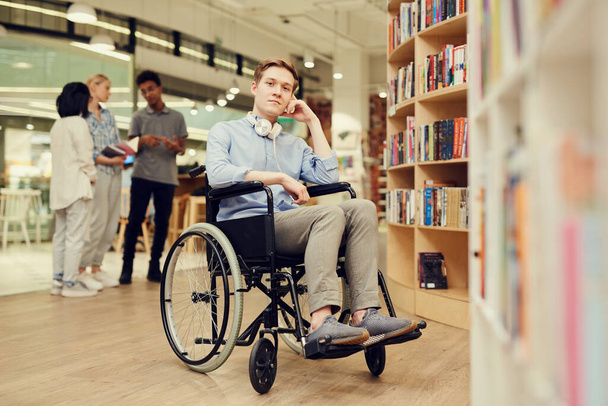 Portrait of content young person with disability student boy with headphones on neck sitting in wheelchair while spending leisure in library - 写真・画像