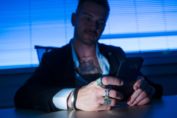 Cagliari, Italia - 20-03-2018: Un blanco rubio con tatuajes vestido con chaqueta y camisa lee los mensajes de su celular sentado en su oficina - Foto, imagen
