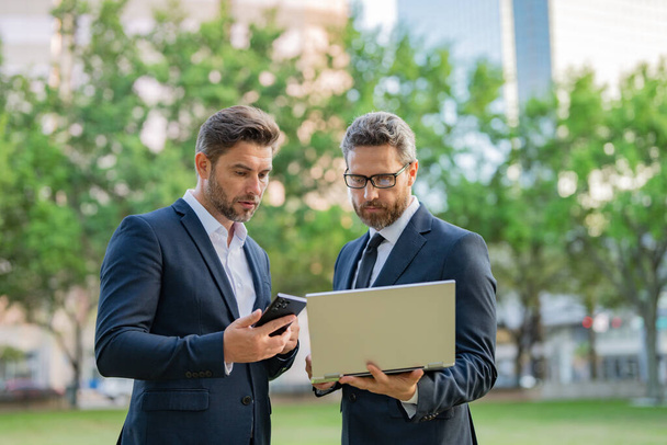 Two handsome young businessmen in classic suits using laptop. Handsome business man in suits working on laptop communication with business projects in city. Partners thinking and plan for business - Foto, Imagen