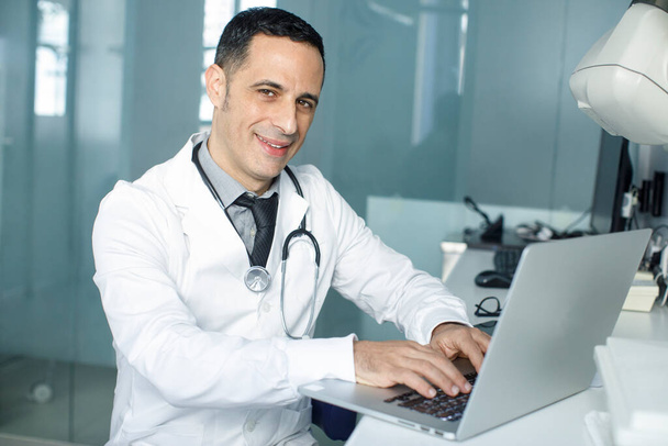 Dunkelhaariger Arzt mit schwarzen Haaren im weißen Mantel, der in seiner Praxis lächelnd am Computer arbeitet - Foto, Bild