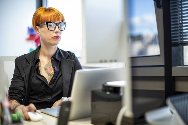 Tattooed woman with red hair and black glasses in a black suit works at her workstation in the office - Фото, зображення