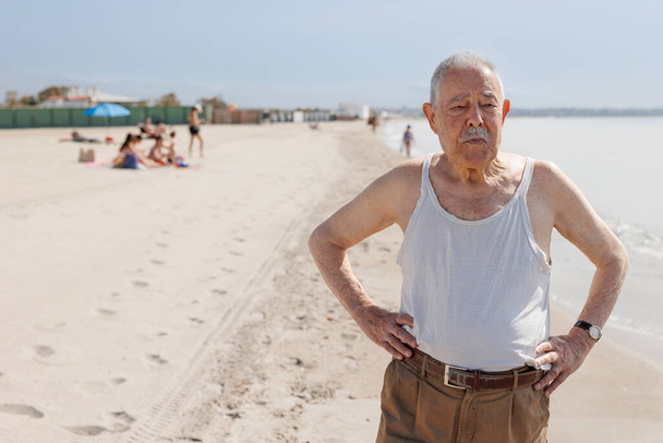 An elderly gentleman dressed in a tank top by the sea is standing with a serious expression - Foto, Imagen