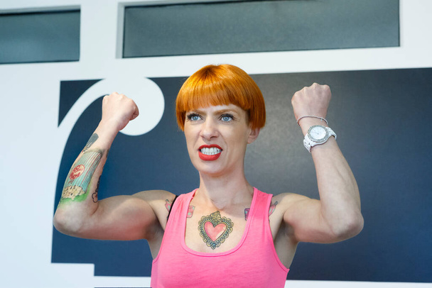 Chica tatuada con el pelo rojo y un top de tanque rosa lanza sus brazos y puños en la ira - Foto, Imagen