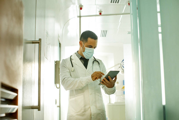 Черноволосый врач в белом халате и галстуке работает, с табличкой стоя в палате больницы - Фото, изображение