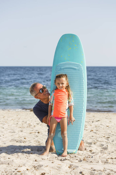 vader met een zonnebril in badpak leert de basis van surfen om zijn dochter op een strand aan zee. - Foto, afbeelding