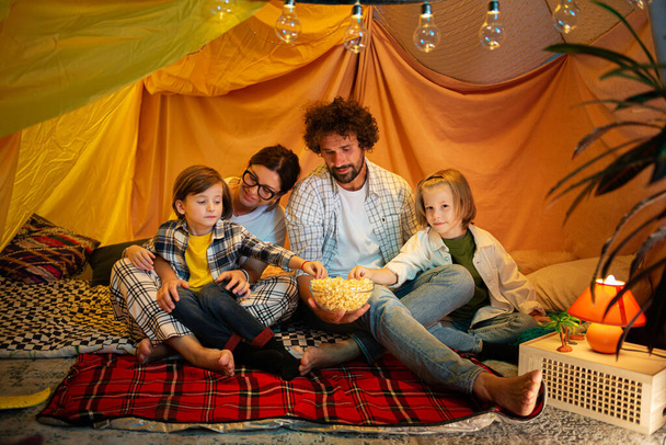 Les parents charismatiques et attrayants passent du temps avec leurs enfants à la maison ils ont fait une grande tente assise à l'intérieur de la tente et regardant un film et mangeant du pop-corn. Famille - Photo, image