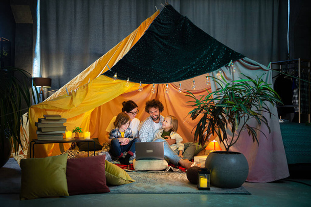 Good time spending with family parents and their two little kids watching movie on the laptop while sitting inside the handmade tent in the kids room. Family - Foto, Imagen