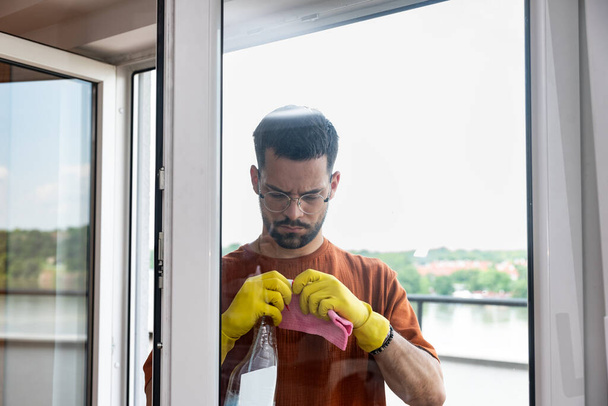 Household and people concept - man in rubber gloves cleaning window with rag and spray cleaner at home. Owner of rental apartment cleaning home for new tenants before they move in as rent agreement. - Фото, зображення
