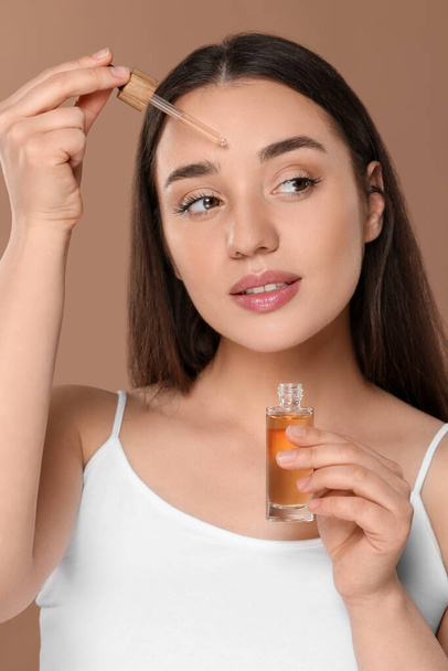 Beautiful woman applying essential oil onto face on brown background - Photo, Image